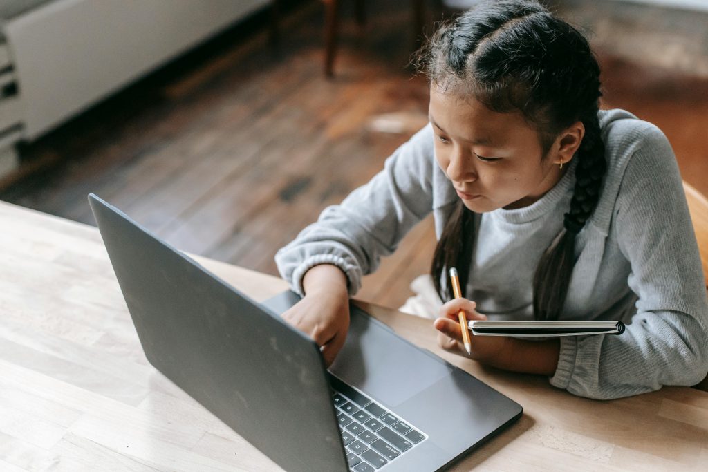 From above of concentrated Asian schoolgirl with pigtails sitting at wooden table with copybook and pencil and browsing laptop for school task